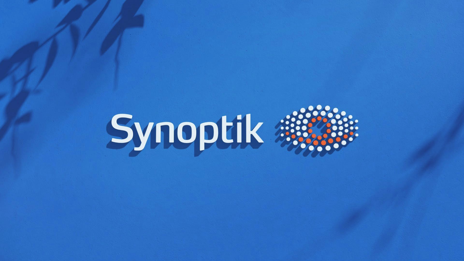  Synoptik-logotypen extrudad från en blå vägg. Skuggor av grenar på väggen.